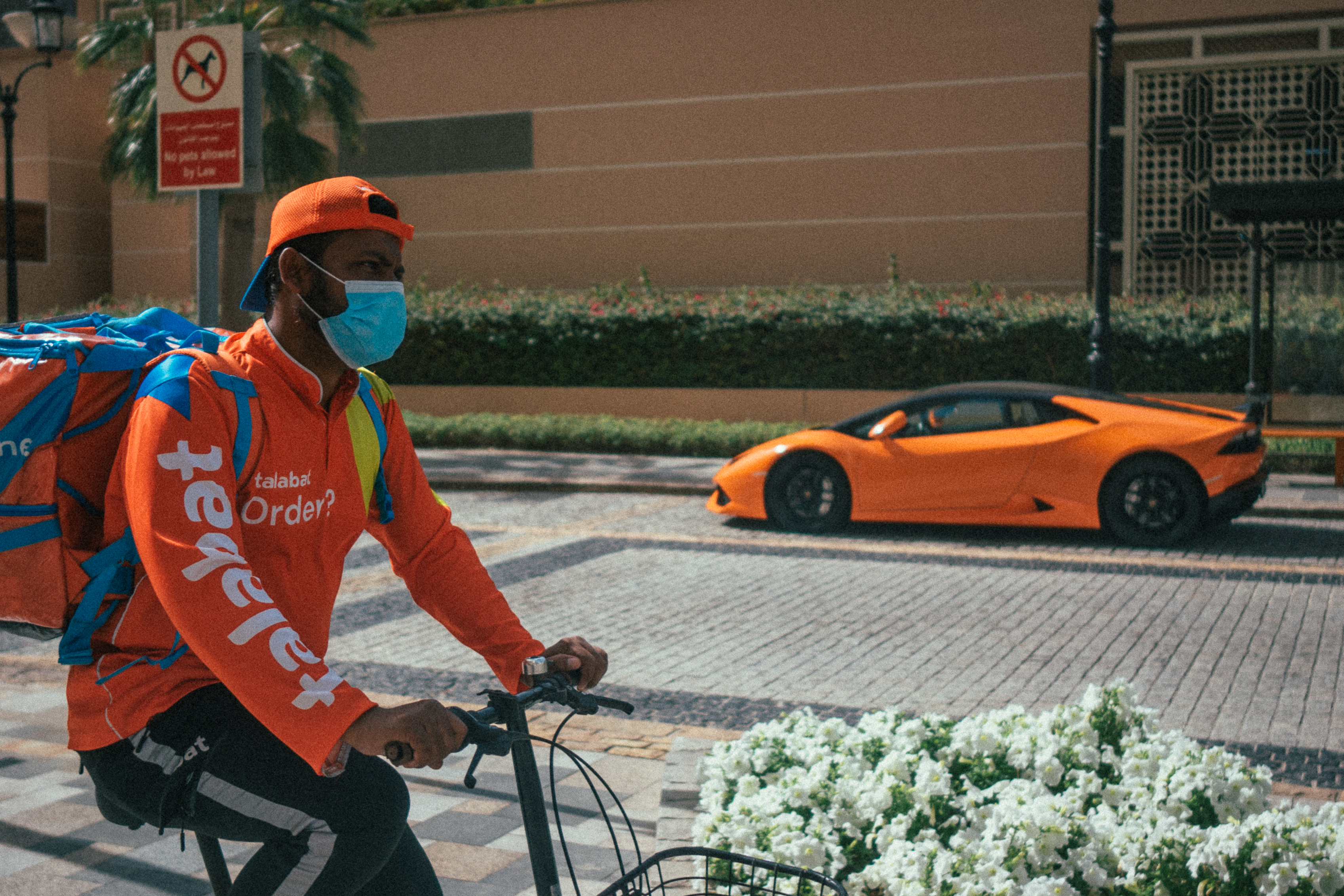 Livreur à vélo au premier plan et voiture de sport orange dans une rue de Dubaï.
