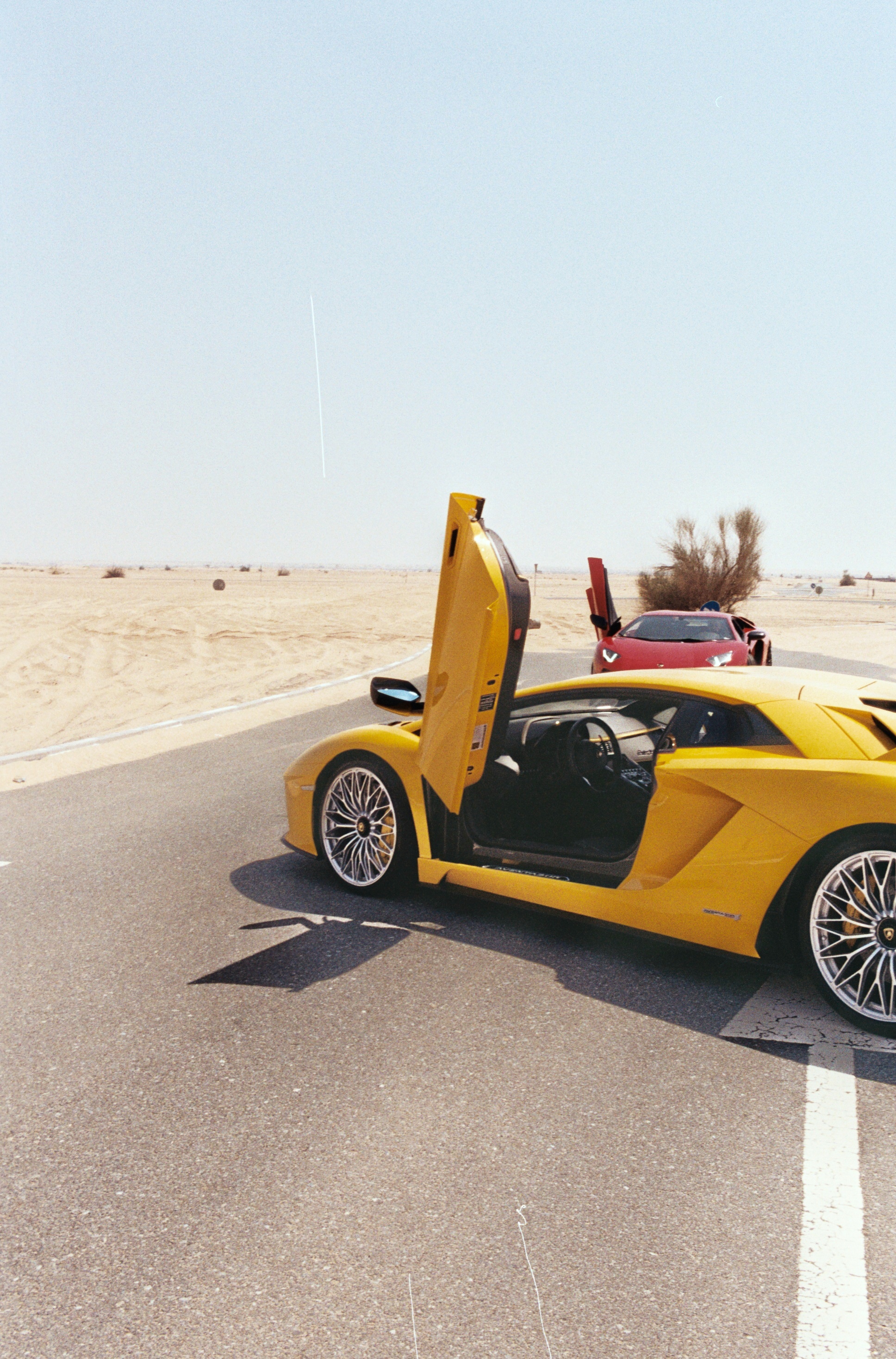 Voiture de sport jaune aux portes relevées sur une route au milieu du sable.
