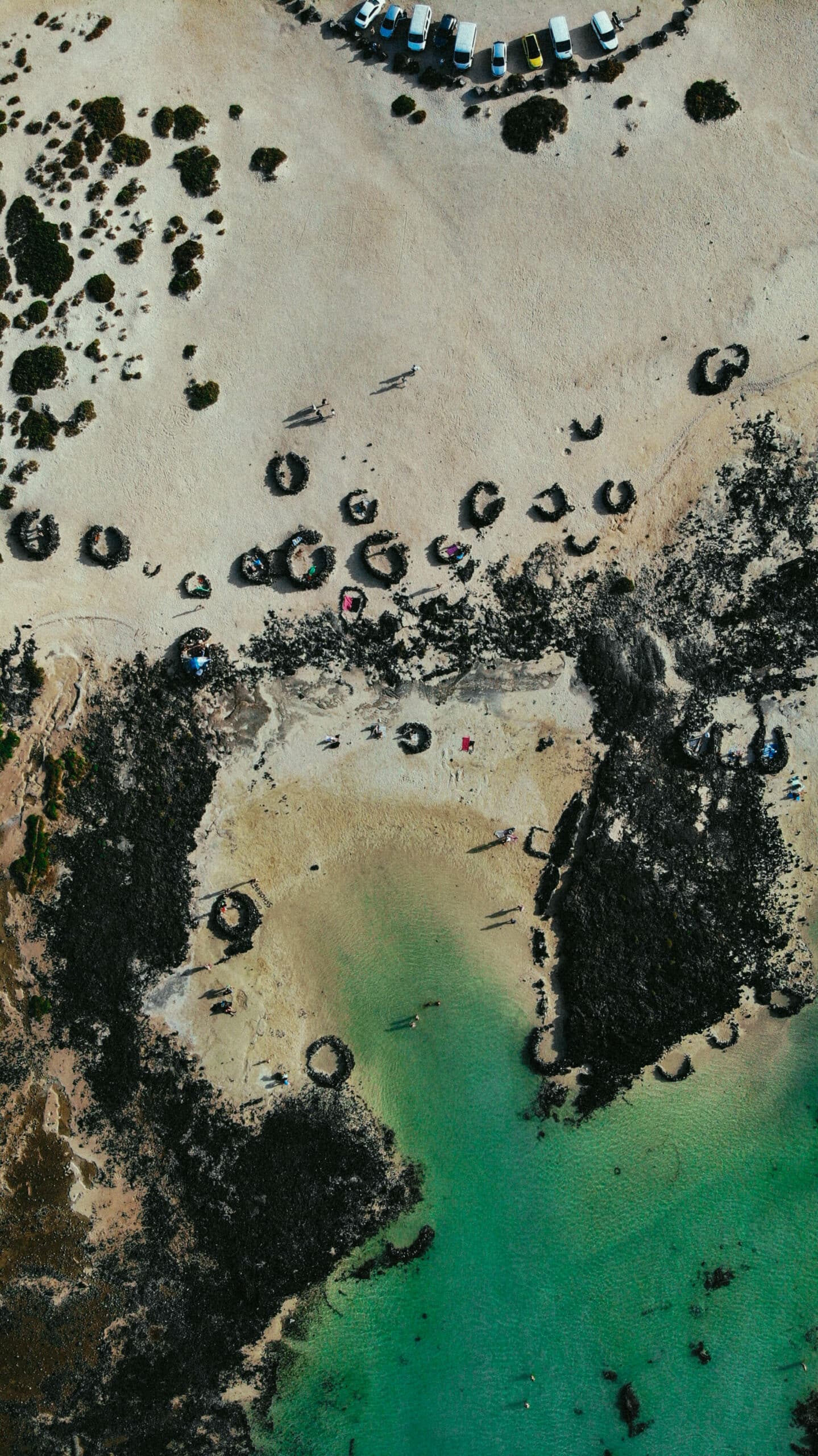 Vue aérienne d’une plage sauvage, avec des eaux turquoise et des abris en pierre.