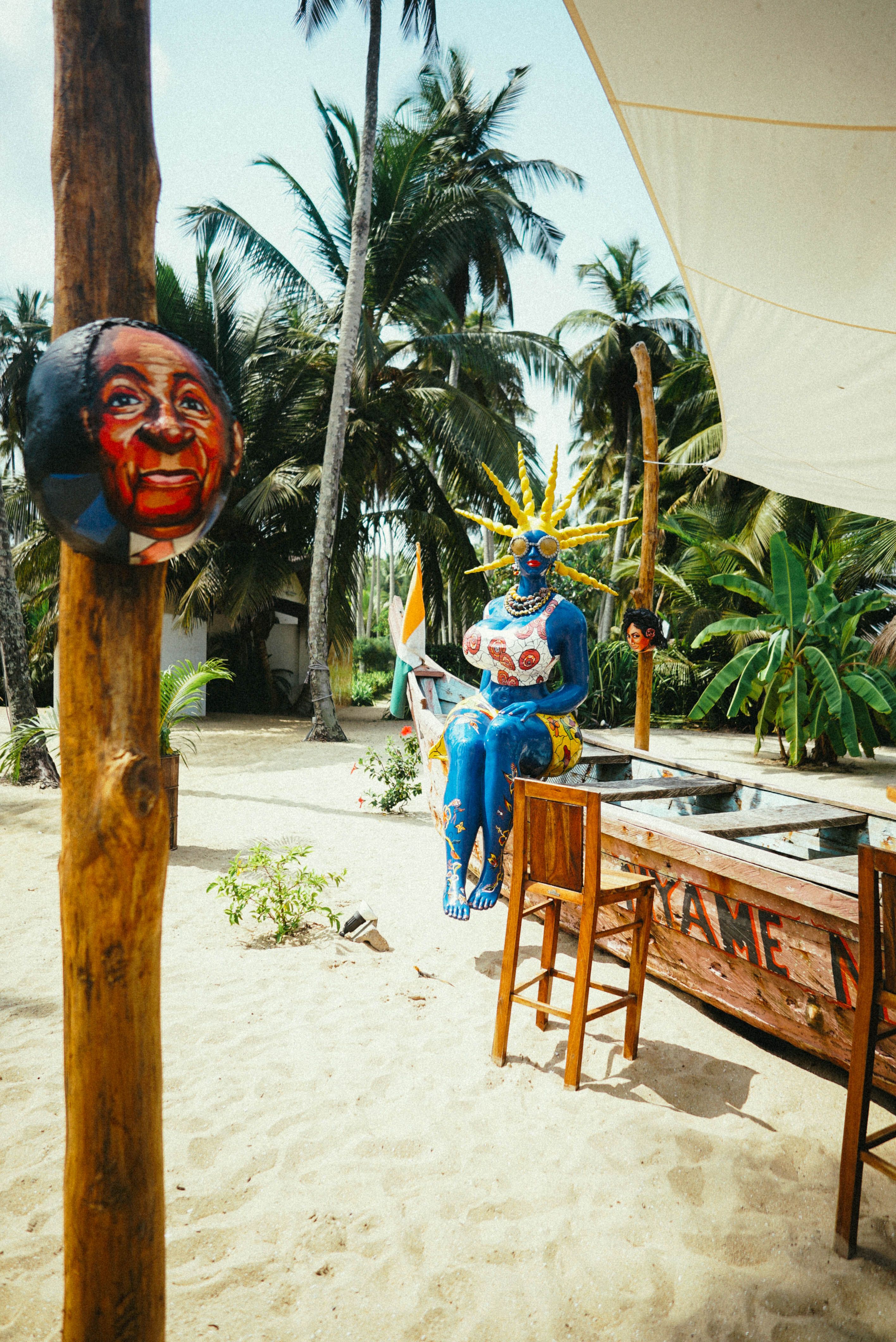 Sculptures colorées, comptoir et tabourets sur le sable sous les palmiers.